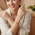 A smiling woman in a cream shirt showcases a 14k gold-filled bracelet with elephant and Figa hand protective charms, indoors by a blurred plant.